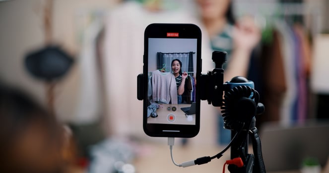 Selective focus on mobile phone of Cheerful woman influencer is live streaming sell clothes on social media or blogging in home studio. Remote buying, Online shopping. photo