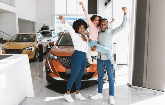 Full length of excited African American family lifting hands up, celebrating new car purchase at auto dealership. Black parents with daughter making YES gesture after buying vehicle at showroom photo
