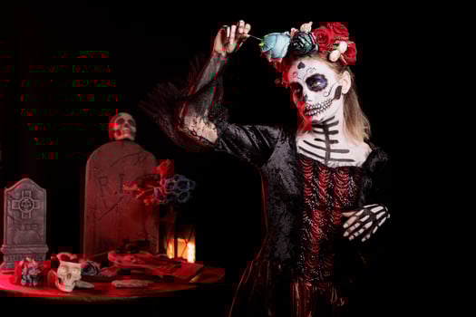 Woman wearing skeleton body paint art, celebrating Day of the Dead. Bizarre lady holding blue rose flower, being surrounded by gothic funeral items during horror Halloween event photo