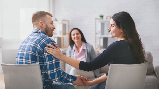 A couple sits facing each other in a therapy session, holding hands. The woman is smiling at her partner, while the man looks happy and relaxed. A lady counselor sits behind them photo