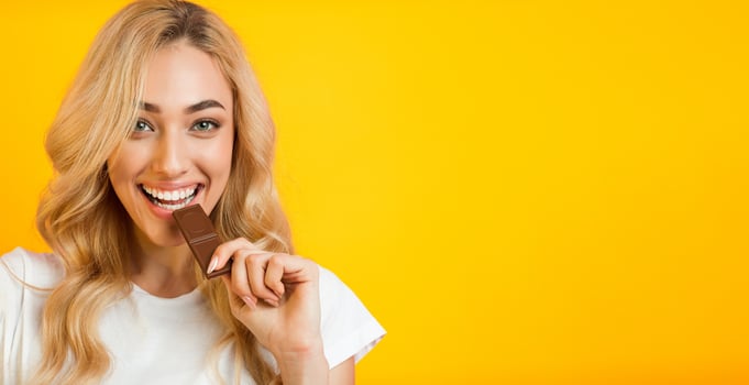 A young woman with long blonde hair smiles while taking a bite of a chocolate bar. She is wearing a white t-shirt and is standing against a bright yellow background. photo