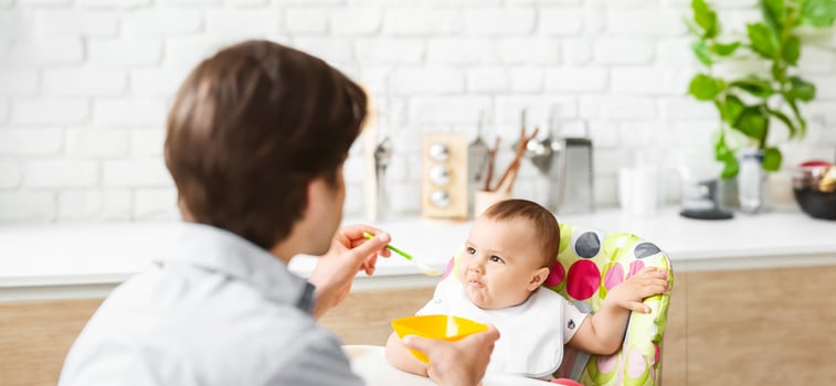 A man feeds a baby in a high chair in a kitchen. The baby is sitting in the high chair, looking at the man who is holding a spoon with food photo