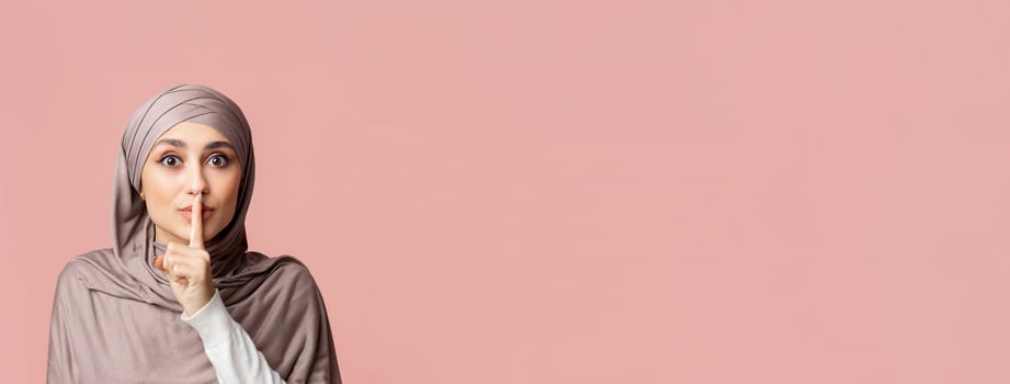 Secret Concept. Young arabic woman in hijab showing hush sign, putting finger on lips, gesturing keep silence over pink studio background, empty space photo