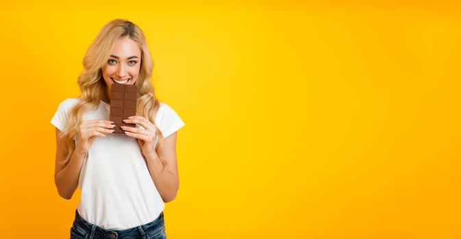 A young blonde woman, wearing a white t-shirt and blue jeans, smiles as she takes a bite from a chocolate bar. The image has a bright yellow background, panorama with copy space photo