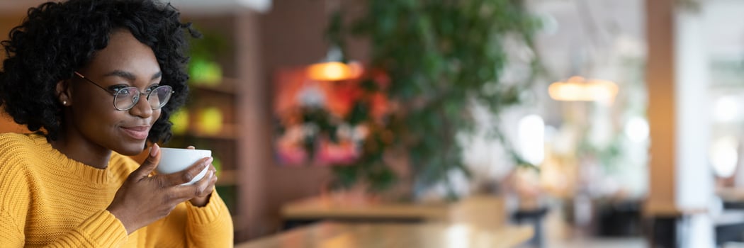 Dreamy young black lady in glasses enjoying cup of coffee in cafe, thinking about future opportunities, close up, copy space photo