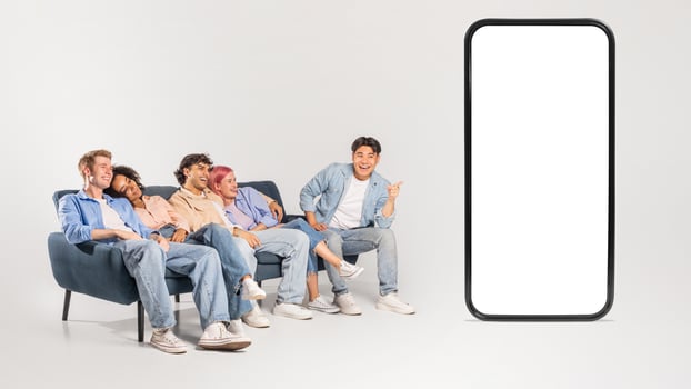 A group of diverse friends, dressed casually in jeans and t-shirts, sit on a blue couch and look at a giant smartphone. One friend points at the blank screen. photo