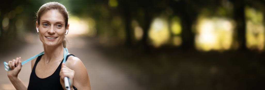 Training equipment. Positive pretty blonde sporty lady in black sportswear exercising at public park, using jumping rope, looking at copy space and smiling, panorama, closeup shot photo
