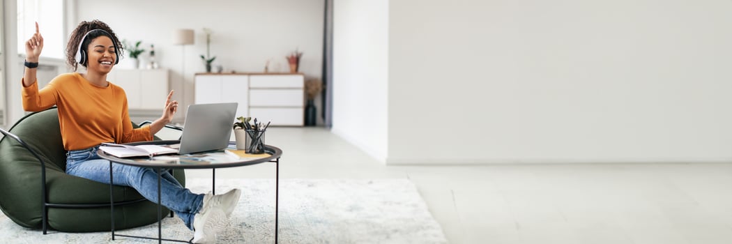 Taking Break From Work. Joyful casual black lady in wireless headphones dancing, having fun and listening to music, sitting on bean bag pouf chair with pc on coffee tea table, free copy space photo
