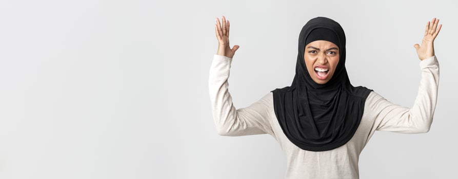 Angry black muslim girl in headscarf shouting and raising hands in fury and looking at camera, posing over light background photo