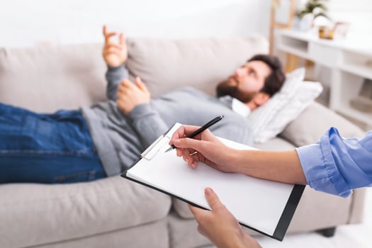 A man is lying on a couch, gesturing with his hand as he speaks, while engaged in a counseling session. A therapist, partially visible in the foreground, is attentively taking notes on a clipboard. photo