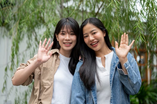 Two beautiful Asian women standing in a green garden, waving their hand and smiling at the camera. photo