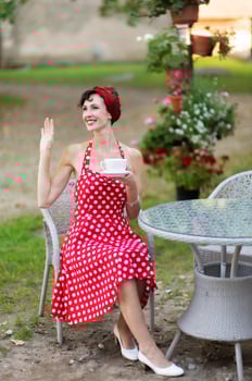 Beautiful smiling woman in red dress with cup of coffee and raised hand in greeting in outdoor cafe photo