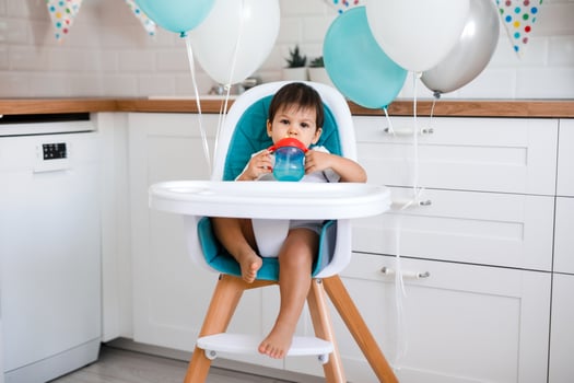 Little baby boy sitting in blue high chair at home on white kitchen and drinking water from sippy cup on background with balloons. photo
