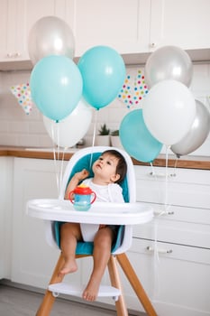 Little baby boy sitting in blue high chair at home on white kitchen and drinking water from sippy cup on background with balloons. photo