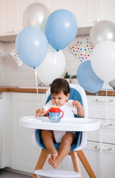 Little baby boy sitting in blue high chair at home on white kitchen and drinking water from sippy cup on background with balloons. photo