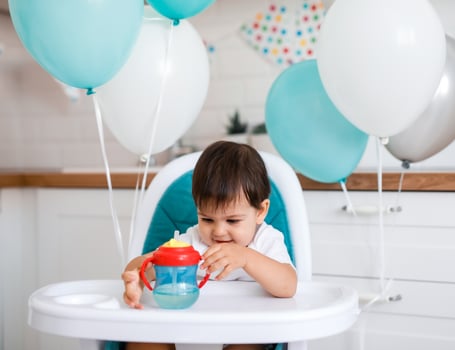 Little baby boy sitting in blue high chair at home on white kitchen and drinking water from sippy cup on background with balloons. photo