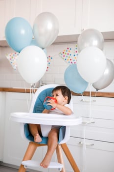 Little baby boy sitting in blue high chair at home on white kitchen and drinking water from sippy cup on background with balloons. photo