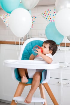 Little baby boy sitting in blue high chair at home on white kitchen and drinking water from sippy cup on background with balloons. photo