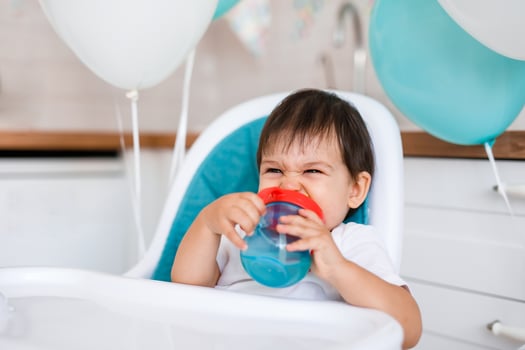Little baby boy sitting in high chair at home on white kitchen and drinking water from sippy cup on background with balloons. photo