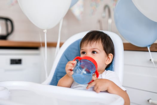 Little baby boy sitting in high chair at home on white kitchen and drinking water from sippy cup on background with balloons. photo