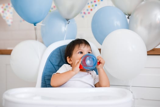 Little baby boy sitting in blue high chair at home on white kitchen and playing with wooden big spoon on background with balloons. photo