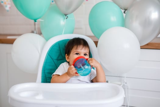 Little baby boy sitting in blue high chair at home on white kitchen and playing with wooden big spoon on background with balloons. photo