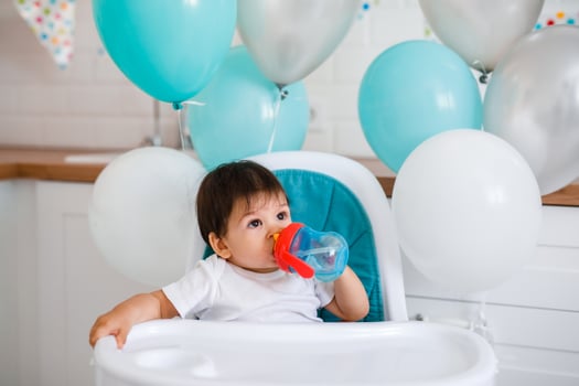 Little baby boy sitting in high chair at home on white kitchen and drinking water from sippy cup on background with balloons. photo