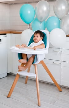 Little baby boy sitting in blue high chair at home on white kitchen and playing with wooden big spoon on background with balloons. photo
