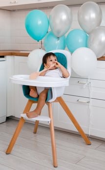 Little baby boy sitting in blue high chair at home on white kitchen and drinking water from sippy cup on background with balloons. photo