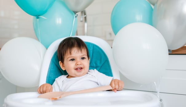 Little baby boy sitting in blue high chair at home on white kitchen and playing with wooden big spoon on background with balloons. photo