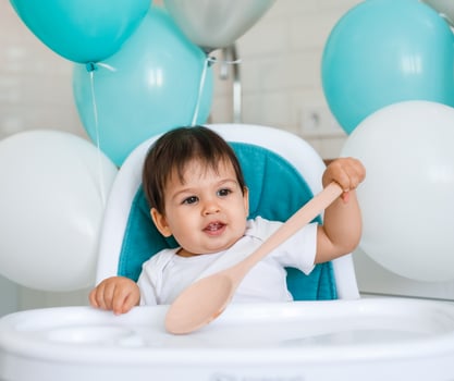 Little baby boy sitting in blue high chair at home on white kitchen and playing with wooden big spoon on background with balloons. photo