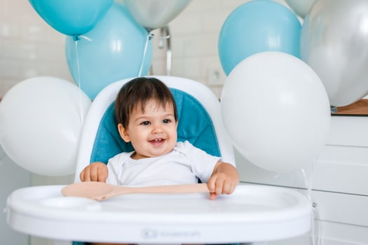 Little baby boy sitting in blue high chair at home on white kitchen and playing with wooden big spoon on background with balloons. photo