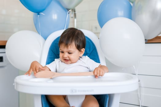 Little baby boy sitting in blue high chair at home on white kitchen and playing with wooden big spoon on background with balloons. photo