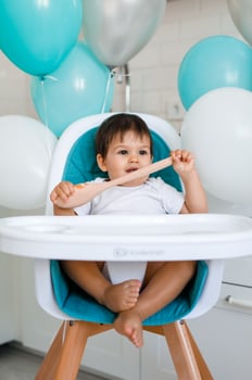 Little baby boy sitting in blue high chair at home on white kitchen and playing with wooden big spoon on background with balloons. photo