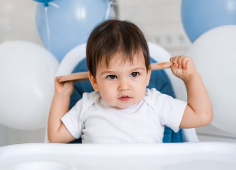 Little baby boy sitting in blue high chair at home on white kitchen and playing with wooden big spoon on background with balloons. photo