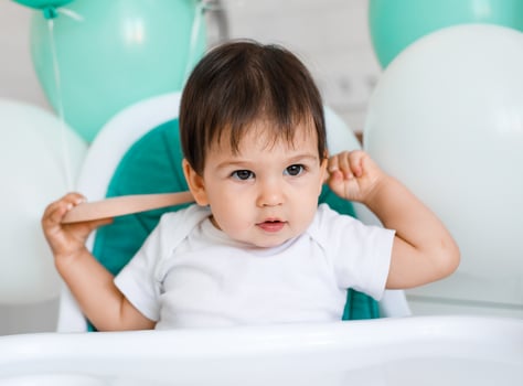 Little baby boy sitting in blue high chair at home on white kitchen and playing with wooden big spoon on background with balloons. photo