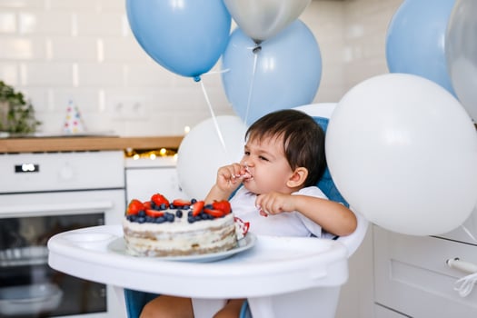 Little baby boy sitting in high chair in white kitchen and tasting first year cake with fruits on background with balloons. photo