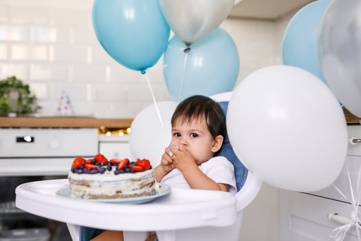 A one-year-old boy sits in a high chair in the kitchen eating birthday cake photo