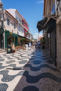 A lively street in Cascais, Portugal, featuring black and white wave-patterned cobblestones, colorful buildings, and people enjoying a sunny day under a clear blue sky. photo