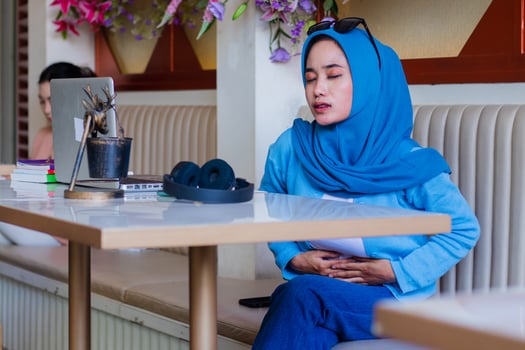 Beautiful Asian woman wearing a veil, covering her eyes, feeling unwell, having a stomachache because of eating the wrong thing, sitting on a sofa in an indoor coffee shop. for advertising, lifestyle photo