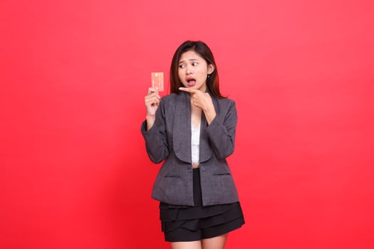Asian office woman's expression shocked candid hand holding a debit credit card while pointing at it wearing a jacket and skirt on a red background. for financial, business and advertising concepts photo