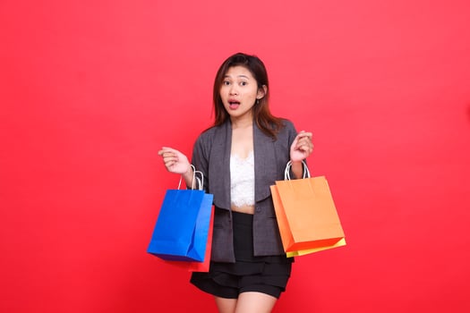 the expression of a shocked Asian businesswoman carrying a shopping paper bag wearing a formal suit on a red background. for commercial, lifestyle and sales concepts photo