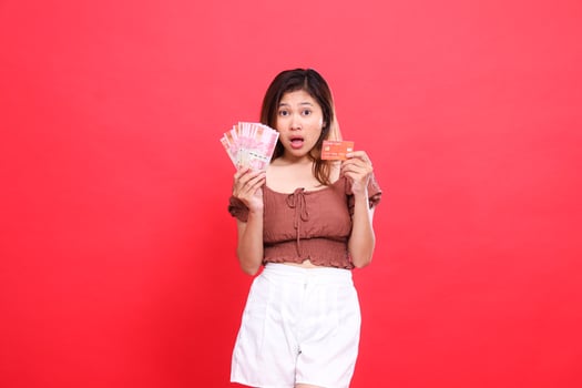 Shocked indonesia woman holding rupiah money and debit credit card wearing brown blouse top with red background. for transaction, technology and advertising concepts photo