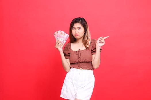 The expression of a sullen Asian girl holding rupiah money while pointing to the left wearing a brown blouse with a red background. for transaction, technology and advertising concepts photo