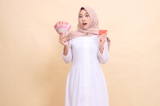 woman indonesia muslim in hijab shocked holding rupiah cash and credit card in hand. Lifestyle, religious, business and finance concepts photo