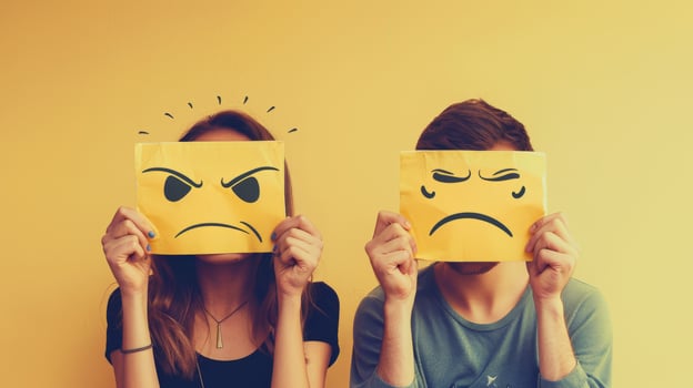 A man and woman hold sad and frustrated emoji cards over their faces in front of bright yellow wall photo