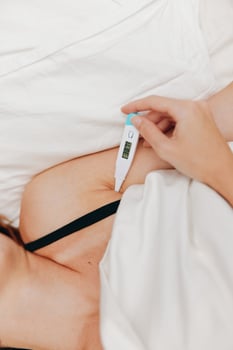 Woman laying in bed, holding an electronic thermometer while another thermometer is placed next to photo