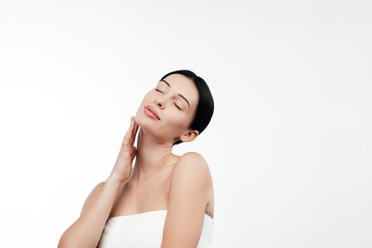 A young woman in a studio against a white background gently touches her flawless skin. Her expression conveys joy and harmony, highlighting her allure and well-groomed appearance photo