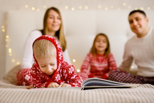 The little boy in festive pajamas is enjoying a book, while his parents and sister can be seen behind him. photo