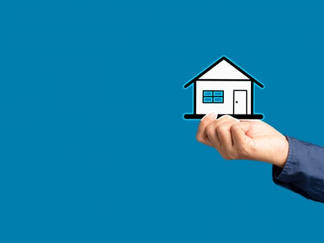 Business and real estate concept. Close-up of hand holding a mini paper house while standing on a blue background. photo